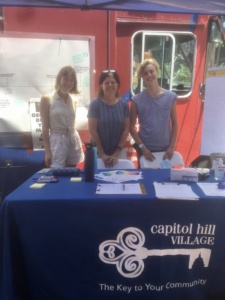 Audrey Barnett, Mary Ellen Curtin, and Kai Walther standing and smiling outside the truck and in front of a table with a "Capitol Hill Village" tablecloth