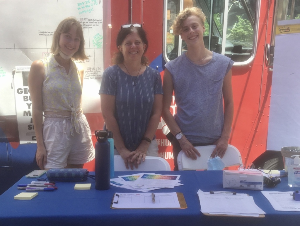 Audrey Barnett, Mary Ellen Curtin, and Kai Walther standing and smiling outside the truck and in front of a table with a "Capitol Hill Village" tablecloth