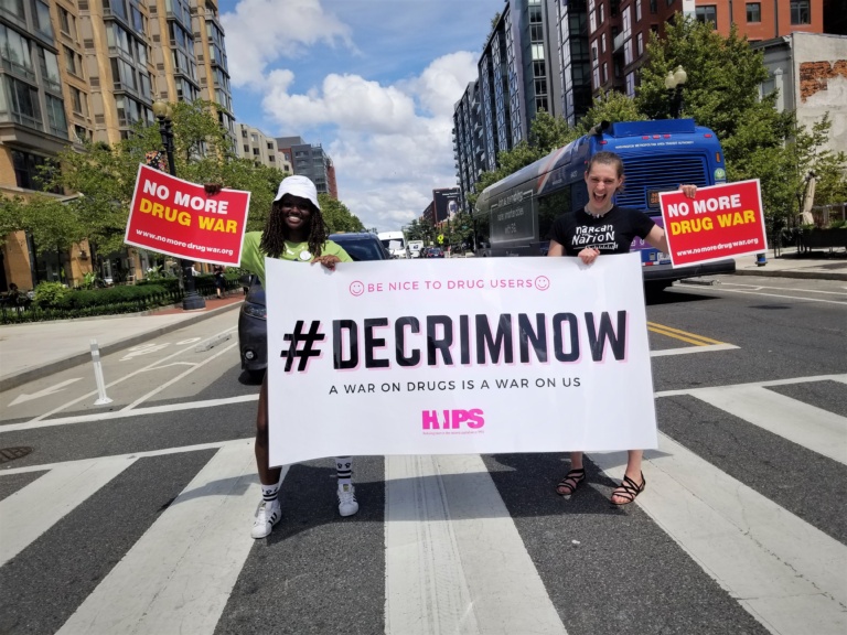 Queen from Drug Policy Alliance (L) and Alex from HIPS (R) holding the banner "#DECRIMNOW" and "NO MORE DRUG WAR" signs. 2021 Photo by Laura Sislen.